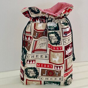 May include: A Christmas-themed drawstring gift bag. The bag features a cream-colored background with rectangular blocks containing holiday phrases like "Merry", "Joy", and "Cheer". The bag's design includes images of reindeer, wreaths, and trucks. The drawstring is dark green, and the interior lining is red and white striped.