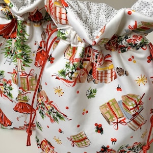 May include: Two white drawstring gift bags with a festive Christmas print. The fabric features illustrations of wrapped presents, women in red dresses, and Christmas trees. The bags have red drawstrings and a silver dotted lining.