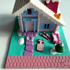 Może przedstawiać: Miniaturowy domek z zabawkami z białym dachem, różowym kominem i jasnoniebieskimi okiennicami. Dom stoi na zielonej podstawie z różową ceglaną obwódką. Akcesoria obejmują sanki, narty i ramkę na zdjęcia.