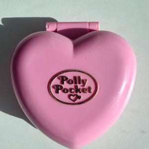 Può includere: Un giocattolo Polly Pocket rosa a forma di cuore con un emblema ovale dorato con la scritta "Polly Pocket" con un simbolo a forma di cuore. Il giocattolo è chiuso e ha un coperchio incernierato. Lo sfondo è di colore azzurro-verde chiaro.
