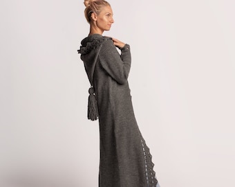 Könnte beinhalten: Ein langer, grauer Strickcardigan mit Kapuze und Quaste. Der Cardigan hat ein Häkeldetail an der Seite.