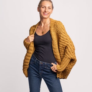 May include: A woman wearing a mustard yellow cardigan with a black vest top and blue jeans. The cardigan has a loose fit and a textured knit.