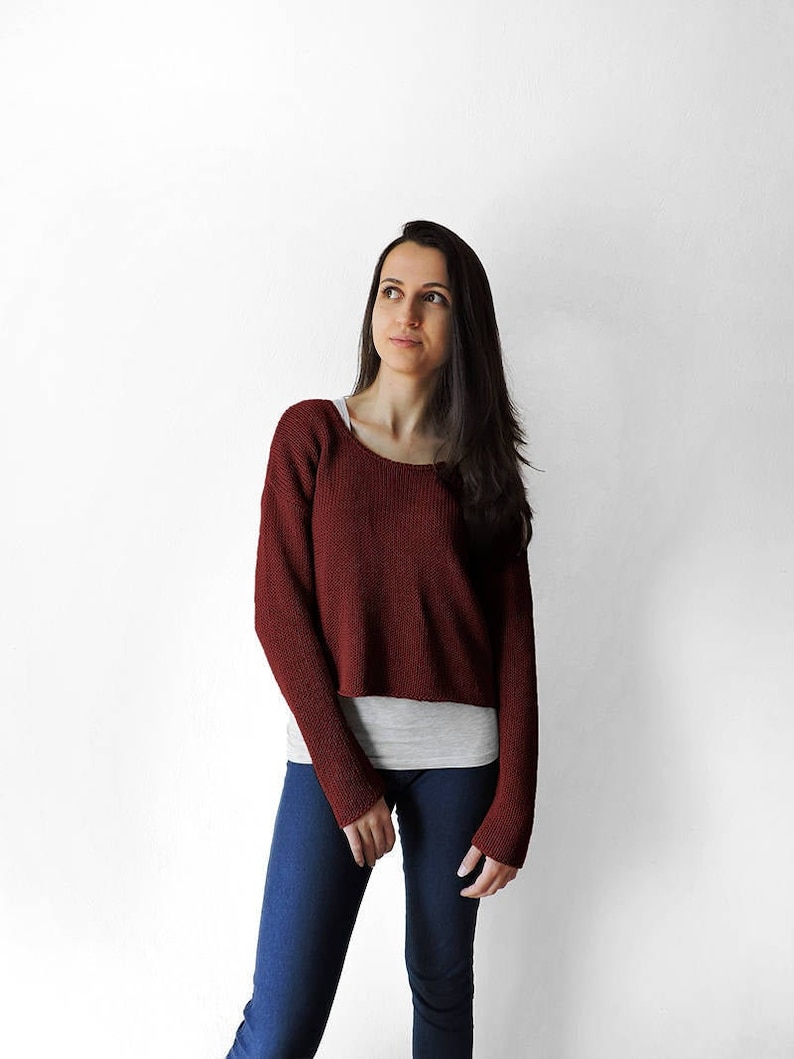 May include: A woman wearing a maroon sweater, blue jeans, and a white undershirt. She is standing in front of a white wall and looking up.