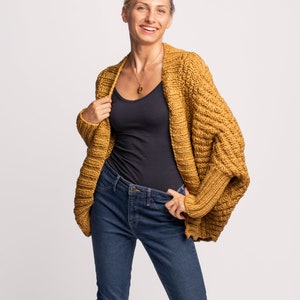 May include: A woman wearing a mustard yellow cardigan with a black vest top and blue jeans. The cardigan has a loose fit and a slightly cropped length.