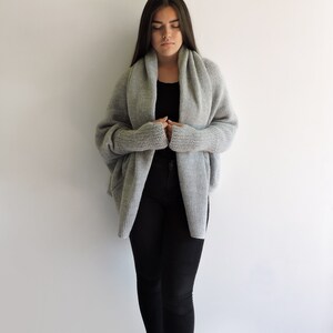 May include: A gray knitted cardigan with a draped collar and long sleeves. The cardigan is worn over a black top and black pants.
