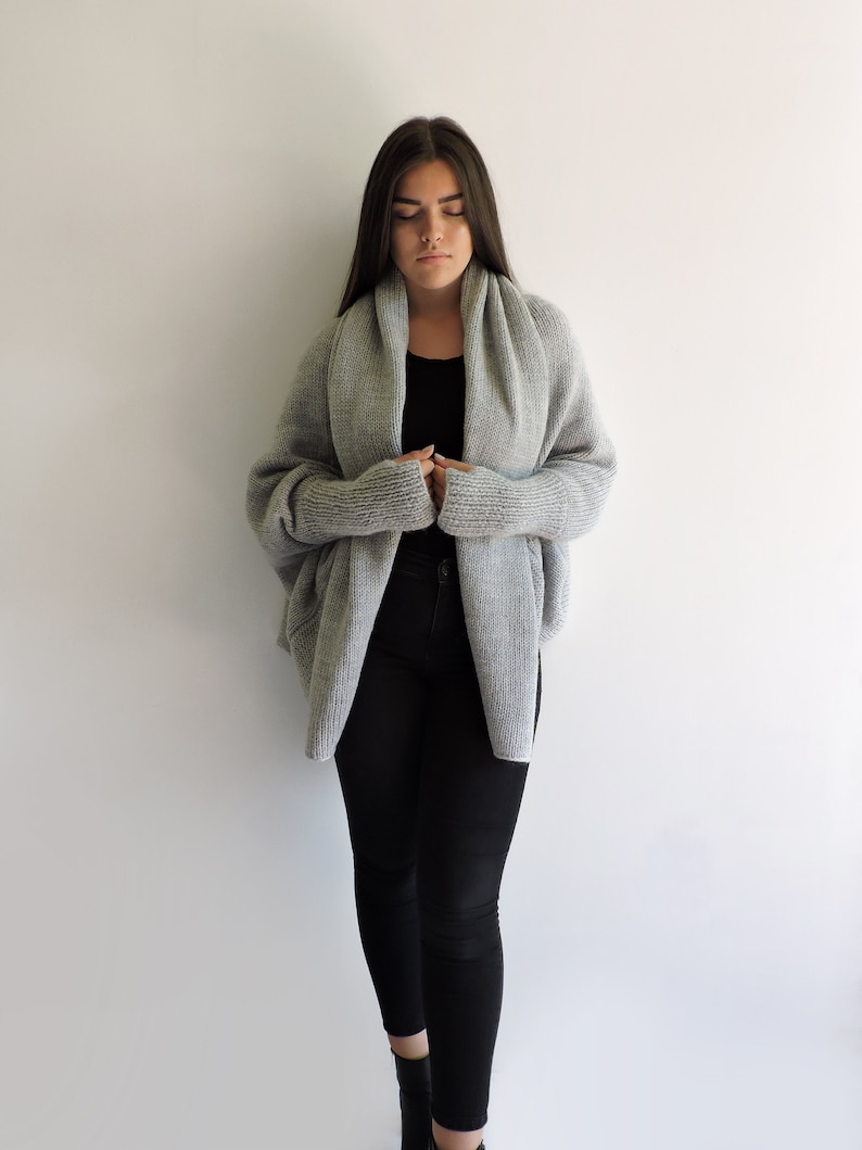 May include: A gray knitted cardigan with a draped collar and long sleeves. The cardigan is worn over a black top and black pants.