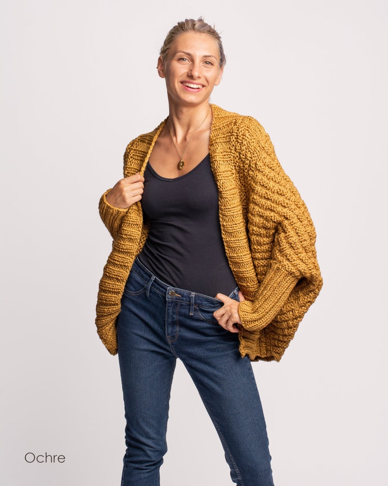 May include: A woman wearing a mustard yellow cardigan with a black vest top and blue jeans. The cardigan has a loose fit and a textured knit.