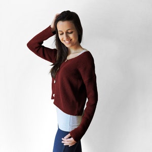 May include: A woman wearing a maroon cropped cardigan with a white undershirt and blue jeans.