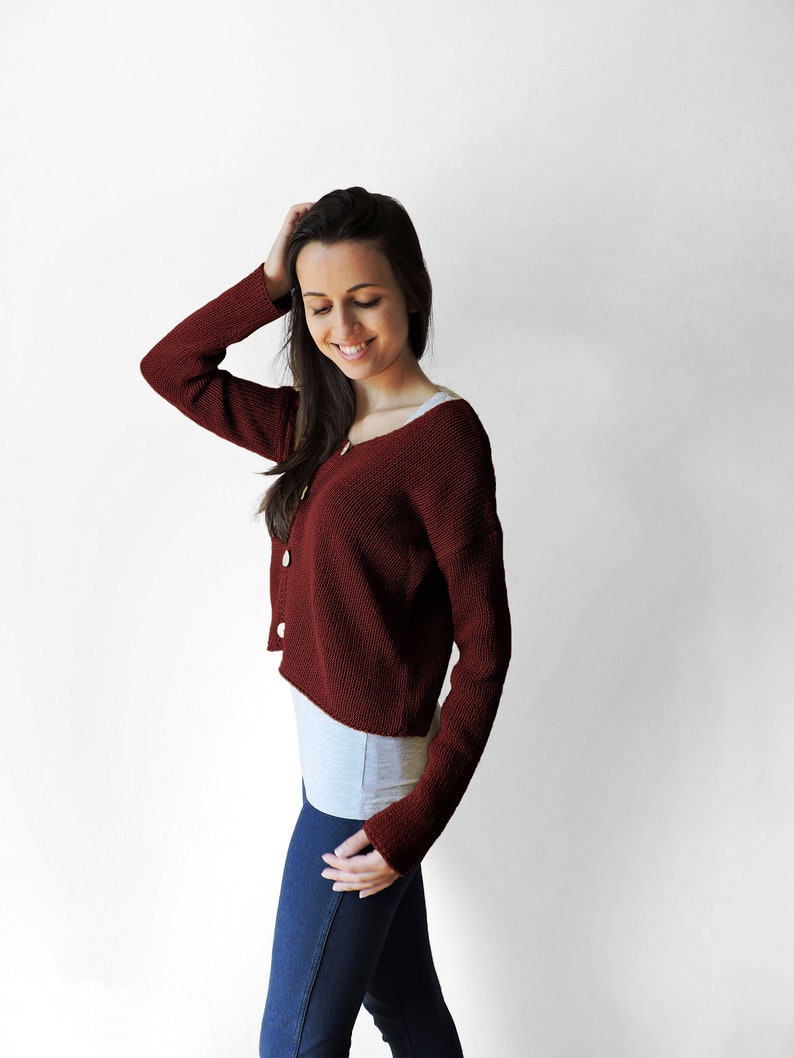 May include: A woman wearing a maroon cropped cardigan with a white undershirt and blue jeans.