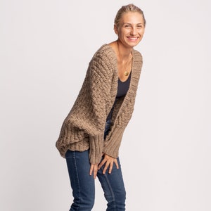 May include: A woman wearing a dark beige knitted cardigan with a relaxed fit and long sleeves. She is also wearing blue jeans.