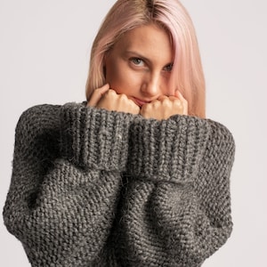Puede incluir: Una mujer con el pelo rosa lleva un su&eacute;ter de punto gris. Est&aacute; sujetando las mangas del su&eacute;ter hasta la cara.