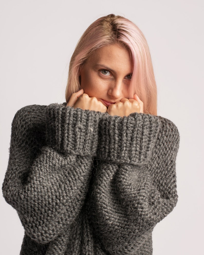 Puede incluir: Una mujer con el pelo rosa lleva un su&eacute;ter de punto gris. Est&aacute; sujetando las mangas del su&eacute;ter hasta la cara.