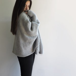 May include: A woman wearing a gray knitted sweater with long sleeves and black pants.
