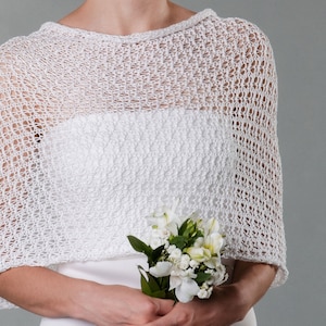 May include: White crochet bridal shawl with a delicate openwork pattern. The shawl is draped over the shoulders of a person holding a bouquet of white flowers.