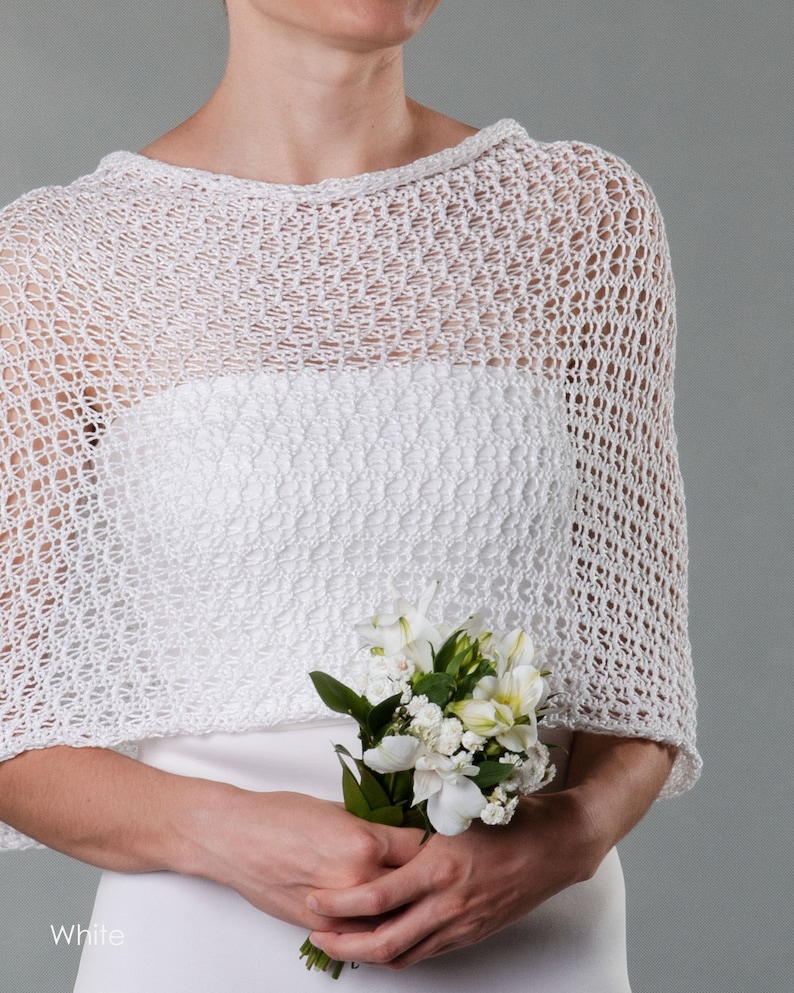 May include: White crochet bridal shawl with a delicate openwork pattern. The shawl is draped over the shoulders of a person holding a bouquet of white flowers.