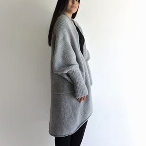 May include: A long, gray, knitted cardigan with a draped, asymmetrical hem. The cardigan has long sleeves and a relaxed fit.