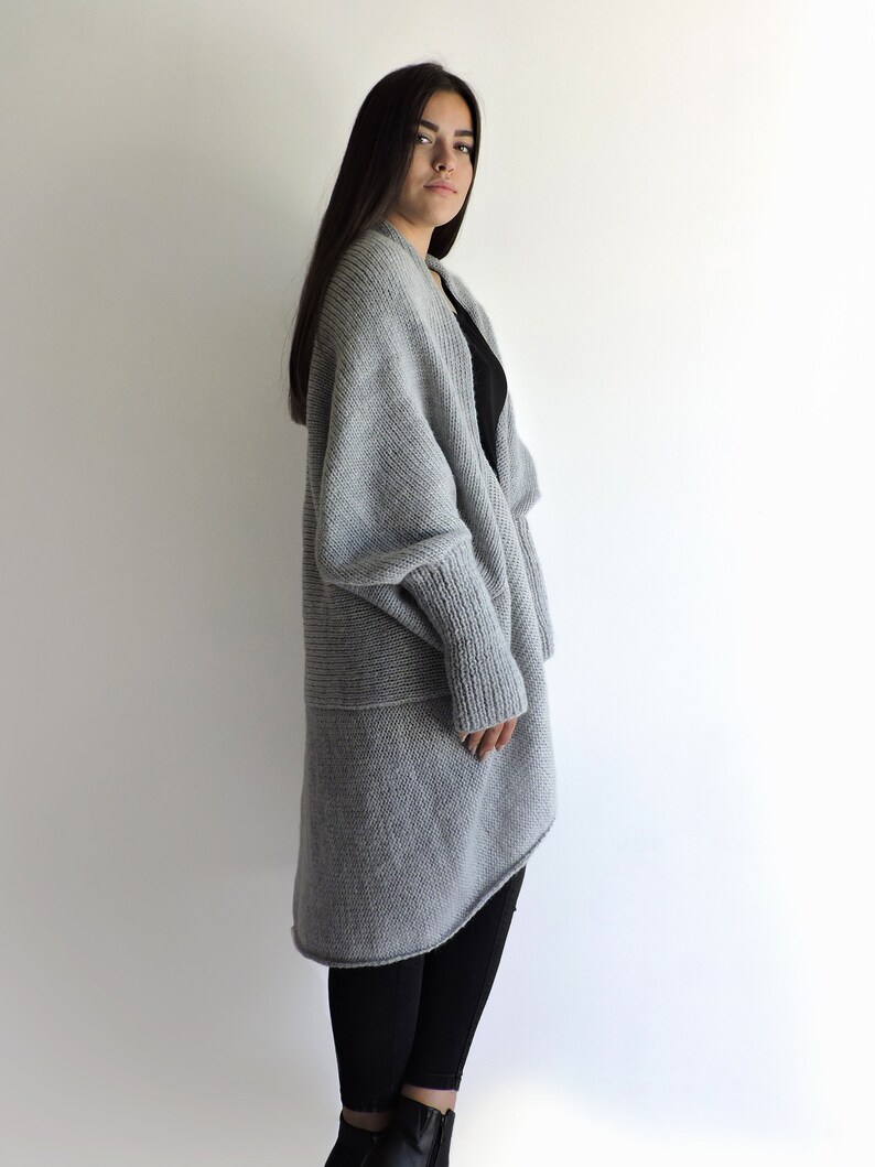 May include: A long, gray, knitted cardigan with a draped, asymmetrical hem. The cardigan has long sleeves and a relaxed fit.