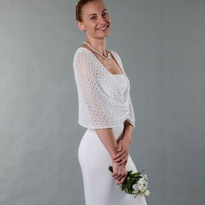 May include: A woman wearing a white crocheted shrug over a white dress, holding a bouquet of white flowers.