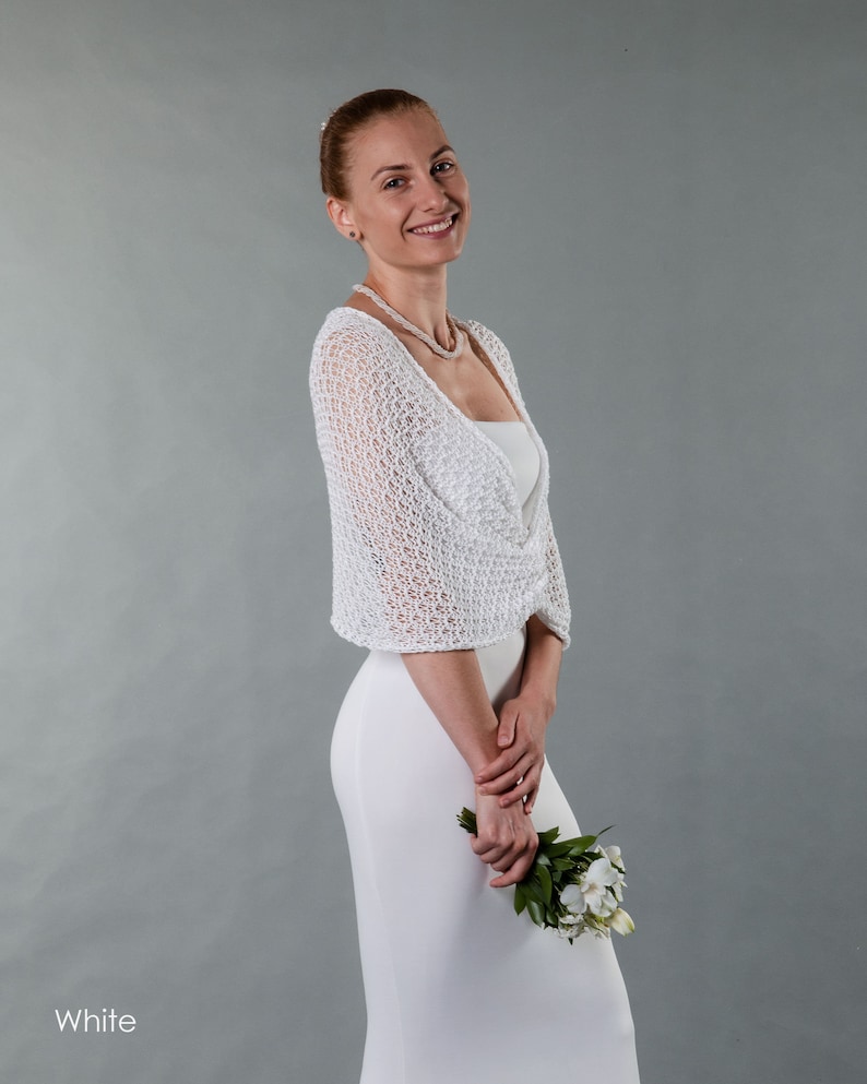 May include: A woman wearing a white crocheted shrug over a white dress, holding a bouquet of white flowers.