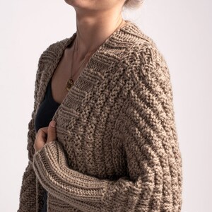 May include: A dark beige knitted cardigan with a cable knit pattern. The cardigan is open at the front and has long sleeves.