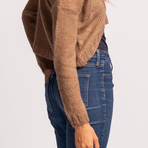 May include: A brown knitted cardigan with long sleeves and a cropped hem. The cardigan is worn over a black tank top and blue jeans.