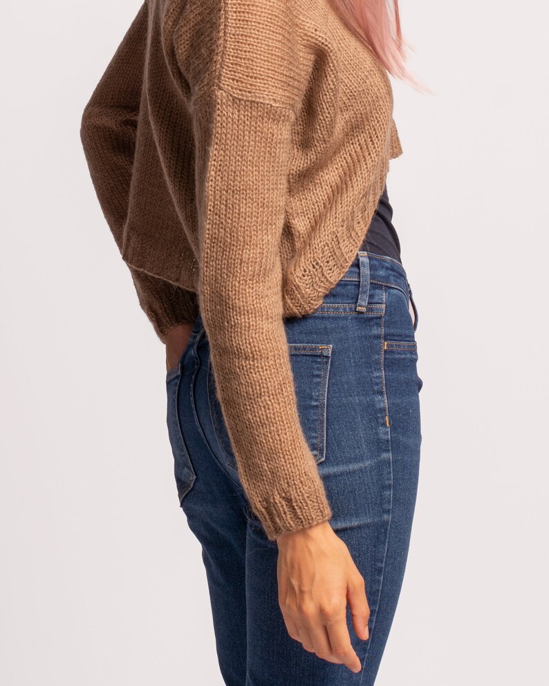 May include: A brown knitted cardigan with long sleeves and a cropped hem. The cardigan is worn over a black tank top and blue jeans.