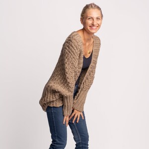 May include: A woman wearing a dark beige knitted cardigan with a relaxed fit and long sleeves. She is also wearing blue jeans.