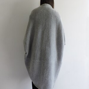 May include: A gray knitted cardigan with a long, loose fit. The cardigan has a ribbed texture and a slightly longer back than front.