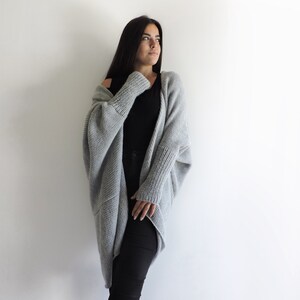 May include: A woman wearing a gray knitted cardigan with long sleeves and a black top. She is standing in front of a white wall.