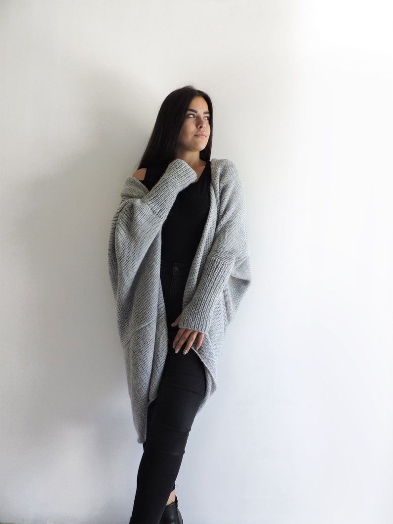 May include: A woman wearing a gray knitted cardigan with long sleeves and a black top. She is standing in front of a white wall.