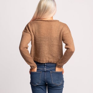 May include: A woman wearing a brown knitted sweater and blue jeans. She is standing with her hands in her pockets.