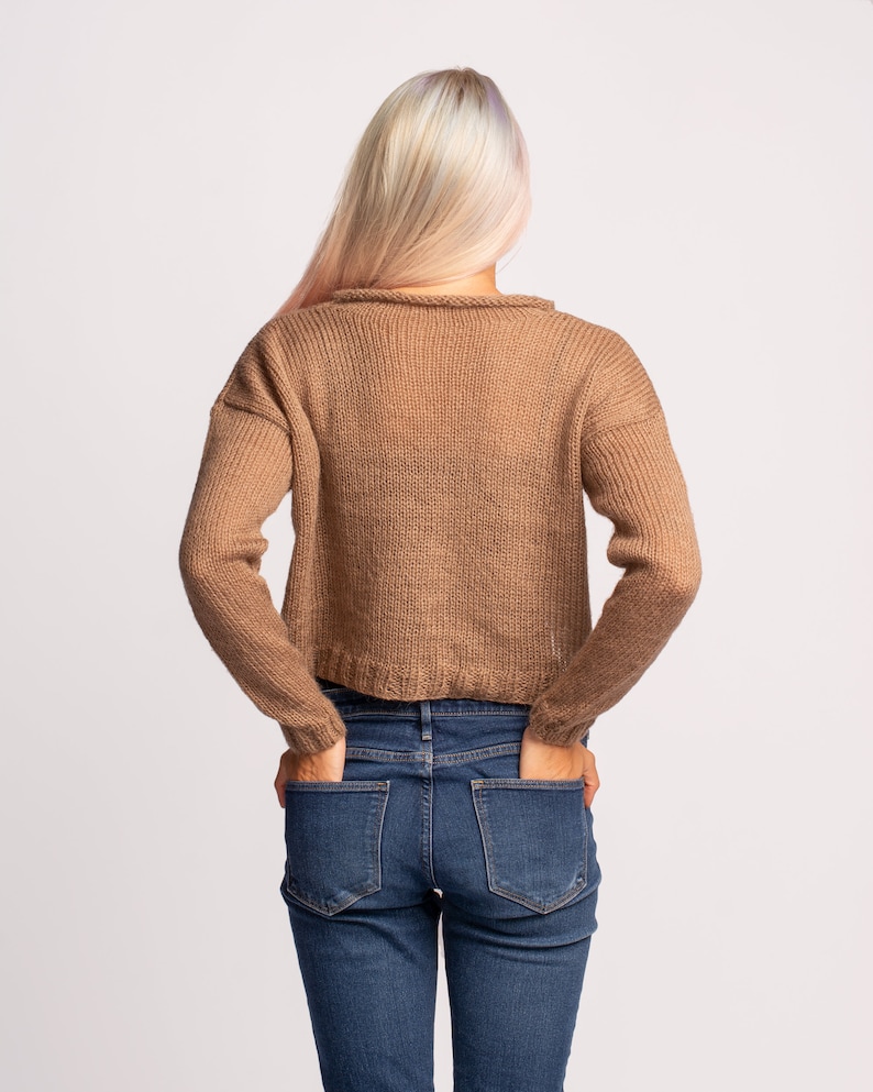 May include: A woman wearing a brown knitted sweater and blue jeans. She is standing with her hands in her pockets.
