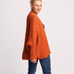 May include: A woman wearing a loose-fitting, orange knitted cardigan with a cable knit pattern. The cardigan has long sleeves and a relaxed fit.