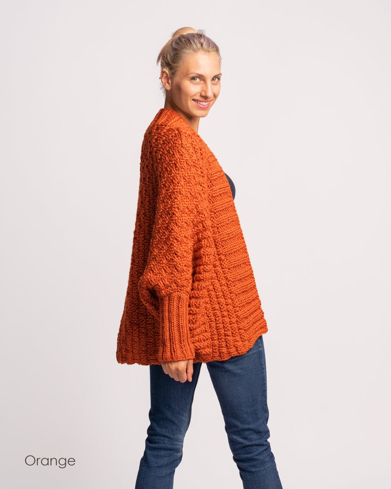 May include: A woman wearing a loose-fitting, orange knitted cardigan with a cable knit pattern. The cardigan has long sleeves and a relaxed fit.
