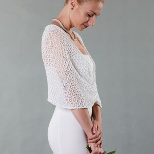 May include: White crochet bridal shrug with a delicate open-weave pattern. The shrug is draped over the shoulders and falls to the elbows. The model is wearing a white dress and holding a bouquet of white flowers.