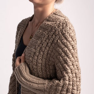 May include: A dark beige knitted cardigan with a cable knit pattern. The cardigan is open at the front and has long sleeves.