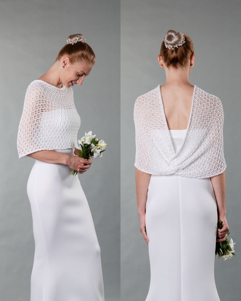 May include: A white crocheted bridal shawl with a delicate openwork pattern. The shawl is draped over a white dress and is worn by a person with their back to the camera.