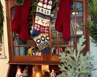 May include: Three Christmas stockings hang from a wooden cabinet. Two are solid red, and one is patterned with snowmen, trees, and reindeer. A small Christmas tree and decorations are on the desk below.