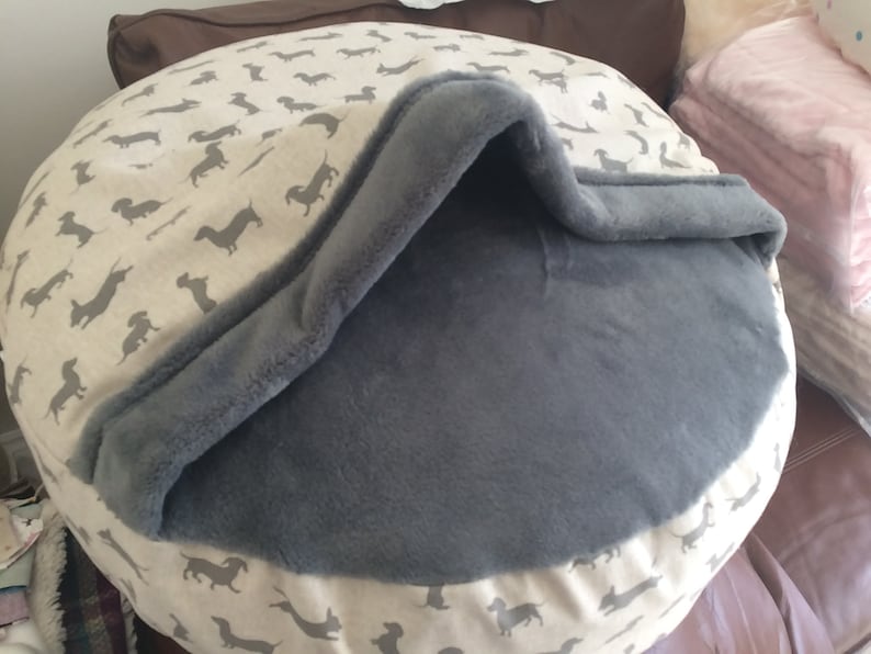 May include: A gray and white dog bed with a removable, gray fleece cover. The bed has a  pattern of small, black dachshund silhouettes on a white background.