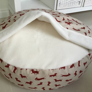 May include: A round, beige dog bed with a white, plush lining. The bed has a red dachshund print.