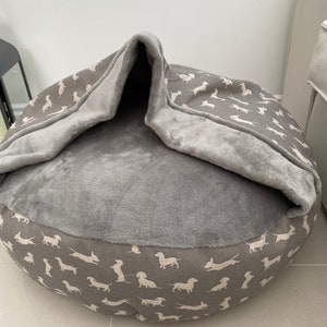 May include: A grey and white dog bed with a removable cover. The cover features a pattern of white dachshunds on a grey background. The bed has a soft, plush lining.