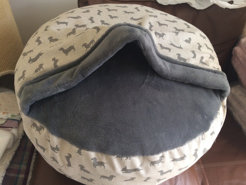 May include: A round, gray and beige dog bed with a removable, gray fleece cover. The bed has a dachshund print on the outer fabric.