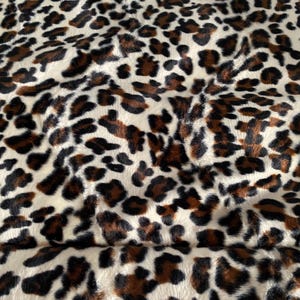 May include: Close-up of a plush fabric with a leopard print pattern. The fabric features a cream-colored background with brown and black spots of varying sizes and shapes, creating a textured appearance.