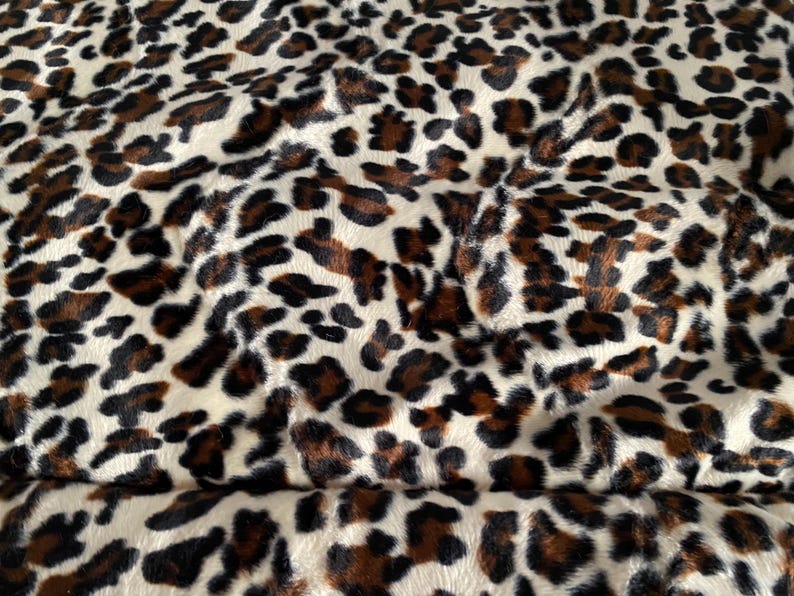 May include: Close-up of a plush fabric with a leopard print pattern. The fabric features a cream-colored background with brown and black spots of varying sizes and shapes, creating a textured appearance.
