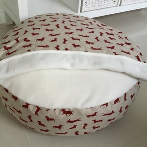 May include: A round beige dog bed with a red dachshund print. The bed has a white fleece lining that can be folded over the top.