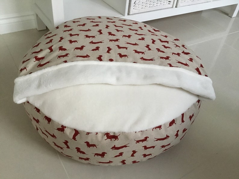 May include: A round beige dog bed with a red dachshund print. The bed has a white fleece lining that can be folded over the top.