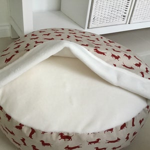 May include: A round beige dog bed with a red dachshund print. The bed has a white fleece lining that can be pulled up to create a cozy den.