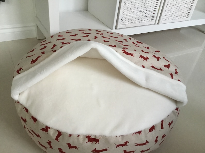 May include: A round beige dog bed with a red dachshund print. The bed has a white fleece lining that can be pulled up to create a cozy den.