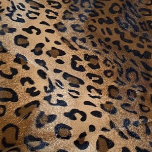 May include: Close-up of a textile with a leopard print pattern. The fabric features a tan background with irregular black and brown spots, creating a naturalistic animal print design. The texture appears soft and possibly plush.