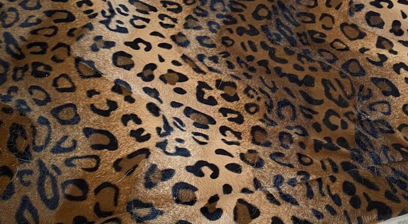May include: Close-up of a textile with a leopard print pattern. The fabric features a tan background with irregular black and brown spots, creating a naturalistic animal print design. The texture appears soft and possibly plush.